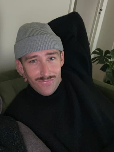 A man with blue eyes and a mustache wears a grey beanie and black sweater while reclining. A plant is visible in the background. The picture is taken at an angle, with the man's face tilted towards the left. He appears to be resting or relaxing.