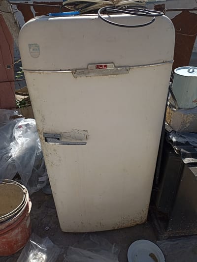 This is an outdoor, eye-level shot of a vintage, off-white refrigerator, likely a washing machine due to its shape. The appliance is weathered and appears to be discarded or stored outdoors. It is positioned in what appears to be a yard or a cluttered outdoor area, with various items scattered around it.

To the left of the machine, there are plastic bags and what looks like a stack of orange and tan buckets. Behind the appliance, a rusty metal structure and some plant life are visible. To the right, a light blue pot with a lid and handles sits on a concrete block, along with other dark, undefined objects that seem to be discarded items. The ground beneath is a mix of cracked pavement and dirt.

The appliance itself has a chrome handle on the front, which is partially covered by tape. There is a circular emblem on the upper left side of the machine with a shield shape and the word "ROSTOCK" printed below it. On the front, above the handle, is a red and silver emblem with Cyrillic text, appearing to read "FROST". The overall condition of the appliance suggests it is no longer in use.

The lighting indicates it is daytime, with sunlight casting some shadows, though no distinct time of day can be determined. The setting appears to be a somewhat neglected outdoor space, possibly a backyard or a storage area, in Sabnova, Russia. No people or specific activities are depicted.