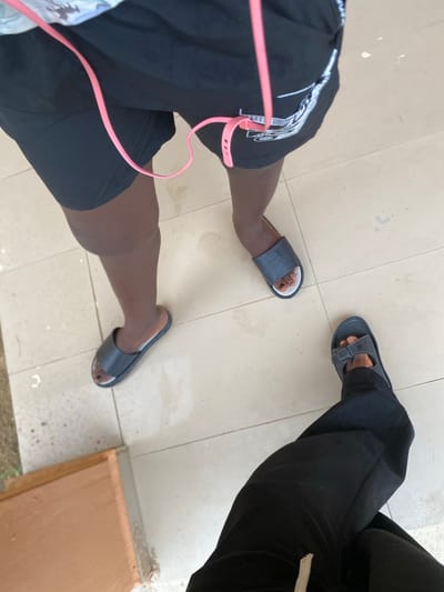 The image shows a top-down view of two people's legs and feet standing on a tiled floor. One person is wearing black shorts with a white graphic pattern and black slide sandals. A pink cord, possibly from headphones, is visible connected to the shorts. The other person is wearing black pants and dark gray slide sandals. The setting appears to be outdoors, with a light-colored tiled floor and some dirt or debris scattered on it. A light brown surface, possibly a wooden plank or a step, is visible to the right of the frame. The lighting suggests it is daytime. The overall impression is casual, with the subjects likely standing or walking. There is no visible text in the image. The location is identified as Dumnga, Nigeria.