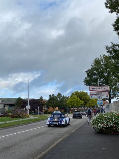 The image depicts a roadside scene in Dorlisheim, France, specifically on a road leading towards Molsheim, as indicated by the prominent directional signage. The sky is largely overcast with grey clouds, though patches of blue sky are visible.

In the foreground and midground, a two-lane asphalt road runs diagonally from the lower left to the upper center. A blue and silver (or white) two-seater convertible car, possibly a replica or classic model, with a rear-mounted spare tire and French license plate "EG-581-MT", occupies the right lane, moving away from the viewer. Behind it, a dark-colored hatchback car (likely a Volkswagen Golf) follows. Several other vehicles are discernible further down the road.

On the right side of the road, a paved sidewalk features a large, manicured flowerbed containing white and red/pink flowers. A group of approximately three to five individuals, dressed casually, walk along this sidewalk in the same direction as the vehicles. A tall, white signpost stands prominently on the right, displaying "MOLSHEIM" and "MOLSE" in red text on white rectangular signs, followed by a smaller sign indicating a twinning agreement. Behind the signpost, a light-colored concrete wall and dense green foliage are visible.

To the left of the road, a central median or roundabout contains a smaller flowerbed with red/purple foliage. In the mid-distance on the left, a tall, grey streetlamp with a modern LED fixture is present. The background features a tree-lined landscape with various buildings partially obscured by vegetation. A distant wooded hill range is visible on the far left horizon, beneath a subtle, faint rainbow arc in the clouds.