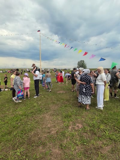 The image captures an outdoor gathering in a grassy field, possibly in or near the village of Кояново, Russia, as indicated by the Russian flag atop a tall wooden pole and a string of triangular, multi-colored flags stretching across the sky. A diverse crowd of people of various ages are present, dressed in casual attire, suggesting a community event or festival. Some individuals are engaged in conversation, while others stand in groups. In the foreground, a group of people are gathered, including a man with a child on his shoulders, and several women. A woman in the center is carrying a shopping bag with "OZON" branding. In the background, more people are scattered across the field, some seated and others standing, observing the proceedings. The environment appears to be rural, with a tree visible in the distance and a cloudy sky overhead.