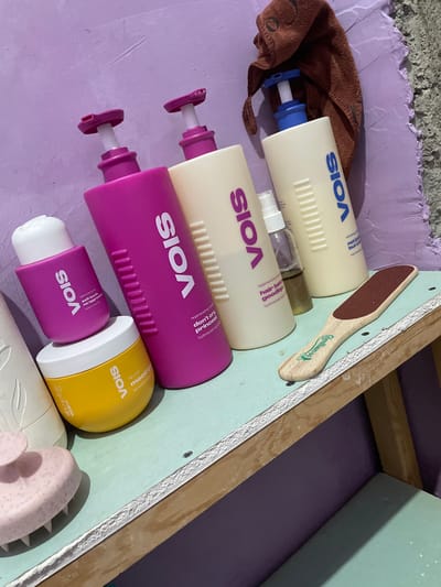 The image shows a collection of VOIS brand personal care products arranged on a purple surface, possibly a shelf or counter, next to a light blue board with a wooden edge. The products include several bottles with pumps and spray nozzles, a yellow container, and a small purple bottle. Text visible on the products includes "VOIS", "mood qu hair mask", "soft touch no aggression", "regenerating cond", and "don't cry princess". A wooden foot file with a red abrasive surface, branded "@millen 60", is also present, positioned on the light blue board. A brown cloth is partially visible in the lower left corner. The overall impression is one of a curated display of beauty or haircare items. The lighting suggests it may be daytime. There is no indication of people or a specific event.