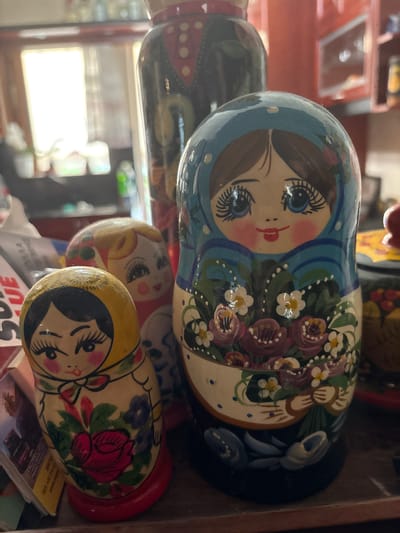 The image shows a collection of Russian nesting dolls, also known as Matryoshka dolls, displayed on a wooden surface. There are at least three dolls visible. The largest doll in the foreground is painted with a blue headscarf, large blue eyes, and is holding a bouquet of flowers, including pink roses and white daisies. To the left and slightly behind it, a smaller doll with blonde hair and a red dress is partially visible. Further to the left and in the foreground, another small doll with a yellow headscarf and a red rose painted on its body is prominently displayed. The background is blurred, but suggests an indoor setting with shelves and objects, possibly in a home or shop. The lighting suggests it is daytime, with light coming from a window. The specific location in Ilion, Greece, is not discernible from the image.