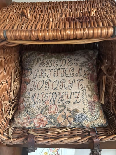 The image displays a woven tapestry-style cushion positioned inside a wicker basket. The cushion, rectangular and plush, features a stitched alphabet from A to Z in uppercase cursive script, arranged in four rows on a light beige background. The letters are dark blue-green. Surrounding the alphabet, and across the bottom of the cushion, is a border of floral and botanical motifs in muted tones of green, rust, light blue, and beige.

The cushion rests within a light brown wicker basket, which appears to be a hamper or trunk given its construction. The interior sides of the basket show additional loose, light-colored straw-like packing material. The upper portion of the image depicts the underside of the basket's lid, also crafted from interwoven wicker. Towards the bottom of the basket, dark brown leather straps with metal buckles are visible, securing the basket's structure or lid. No people are present. The specific location within the city of Monterey, United States of America, is not discernible from the visual information provided in the image.