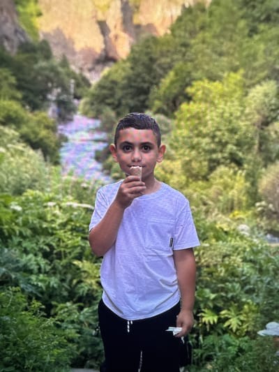 A young boy stands in a lush green outdoor environment, holding a small ice cream cone to his mouth. He is wearing a white t-shirt and black sweatpants. The background features abundant green foliage and hints of rocky terrain, suggesting a natural setting possibly within or near the city of Artik, Armenia, a region known for its scenic landscapes.