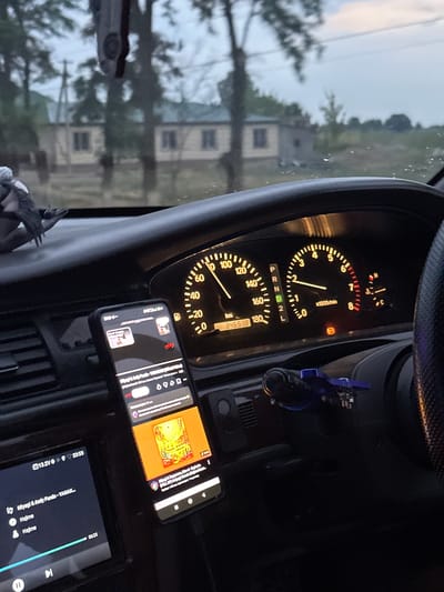 The image captures the interior of a car, likely in Достук, Kyrgyzstan, viewed from the driver's perspective. The time of day appears to be dusk, with a soft, diffused light suggesting an overcast sky or the early evening.
Dominating the foreground is the car's dashboard. A prominent speedometer displays a speed of approximately 40 km/h, with the odometer reading 346518 km. Next to it, a tachometer shows the engine RPM is around 1000. Various warning lights are visible, including a low fuel indicator and an engine warning light.
Two smartphones are mounted on the dashboard. The phone on the right is displaying album art for a music track titled "Miyagi & Andy Panda - YAMAK...". The second phone, partially obscured, shows a music interface with song titles like "Hajime".
Through the car's windshield, a blurry landscape is visible, featuring buildings that appear to be houses or apartments, set against a backdrop of trees and a muted sky. The overall scene evokes a sense of a casual drive, possibly during a commute or a journey, with music playing in the background. The ambient lighting and the interior view suggest a personal and functional space within the vehicle.