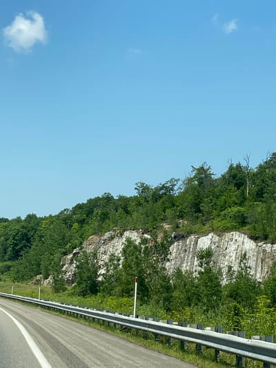 The image shows a roadside view in Pointe-au-Chêne, Canada, on a clear, sunny day. A paved road curves to the left, with a metal guardrail running alongside it. Beyond the guardrail is a grassy area dotted with small plants and wildflowers, leading to a steep, rocky cliff face. The cliff is predominantly light gray and white with distinct vertical striations, indicating a geological formation. Lush green trees and various shrubs cover the top of the cliff and extend down its slopes. The sky is a vibrant blue with a single, small white cloud positioned in the upper left quadrant. No people or vehicles are visible in the scene.