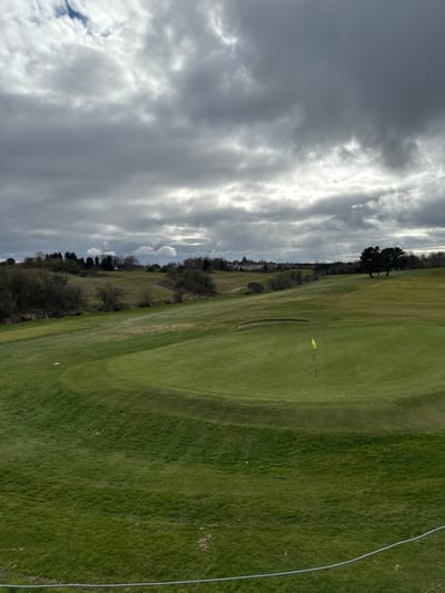 The image depicts a golf course, likely the Pitreavie Golf Club, located in Dunfermline, United Kingdom. The foreground consists of well-maintained green grass, likely part of a fairway or rough. A yellow flag marks the location of a hole on the green. A sand bunker is situated near the green. Beyond the green, there's a line of trees and bushes separating the golf course from an area with some houses or buildings in the distance. The sky is overcast with heavy, textured clouds. A silver fence is running the right side of the image.