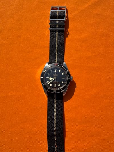 A Tudor Black Bay Fifty-Eight watch is centered on a dark-striped NATO strap with a lighter stripe, on a bright orange textured surface. The watch has a black dial and bezel with a silver case. The strap buckle is on the left side of the frame. The scene's location cannot be determined based solely on the image and information provided.
