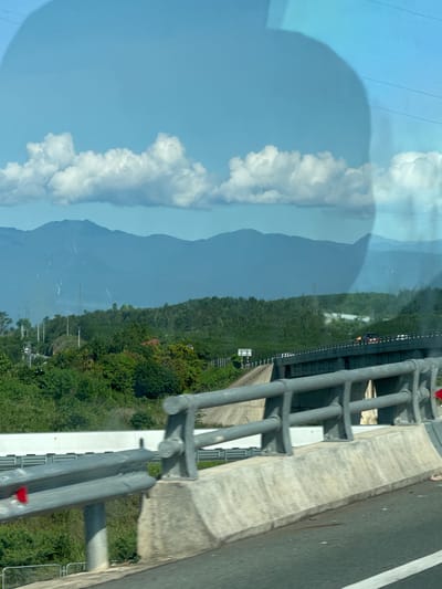 The image depicts a scenic view of mountains in the distance, with lush green foliage and trees in the mid-ground. A highway bridge with guardrails is prominently featured in the foreground, suggesting the viewpoint is from a vehicle traveling on or alongside the road. The sky is a clear blue with scattered white clouds. The scene is likely captured in Dien Tho, Vietnam, given the typical landscape of mountainous terrain and abundant vegetation in such regions. There are no people or direct interactions visible in the frame. The environment is characterized by natural beauty with man-made infrastructure like the highway bridge.