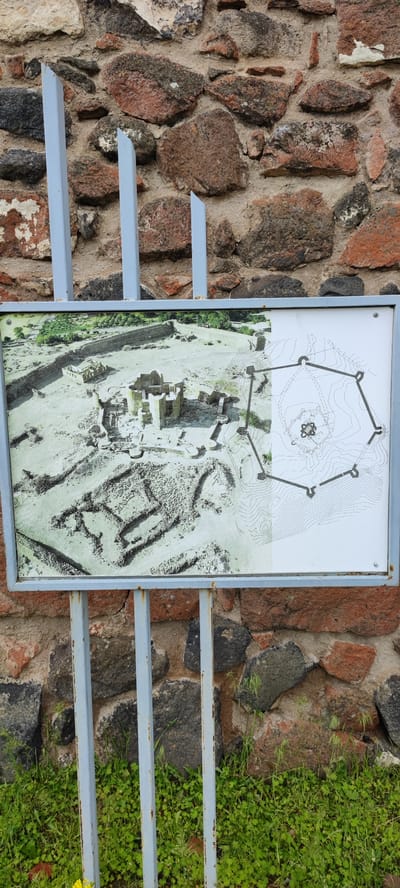 The image shows a signpost in front of a stone wall in Dashtadem, Armenia. The signpost, framed in grey metal, presents information about a historical site, with an aerial view photograph and a schematic map displayed side-by-side. The photograph shows the ruins of a fortress or ancient structure, revealing walls, towers, and other architectural elements. The map illustrates the layout of the same site, outlining the perimeter and key features with contour lines indicating the topography. The wall behind the signpost is constructed from irregularly shaped stones of varying sizes and colors, predominantly shades of brown, grey, and red. In the foreground, green vegetation, including grass and small flowering plants, grows at the base of the wall and around the supporting metal posts of the sign. The scene is outdoors, likely in an area near the historical site represented on the sign.