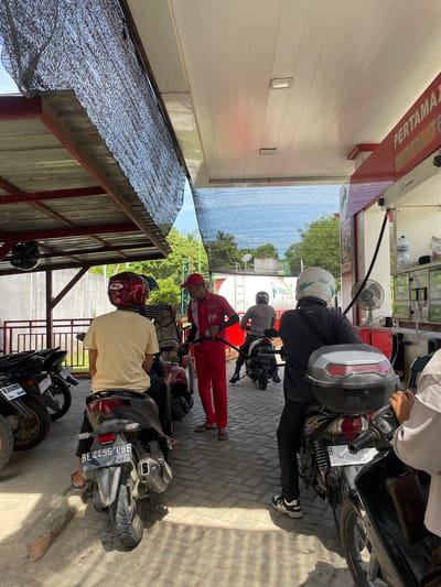 The image depicts a scene at a gas station in Glee Gogo, Indonesia. Several people on motorcycles are waiting to refuel. A gas station attendant, wearing a red uniform and cap, is seen holding a gas nozzle and attending to a motorcycle. The background shows a white building with "PERTAMAX" signage, indicating it is a Pertamina gas station. Trees and a blue sky are visible in the distance. The ground is paved with light-colored bricks.