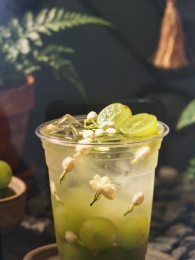 The image shows a close-up of a clear plastic cup filled with a pale green beverage, ice cubes, and garnished with what appear to be small white flowers and slices of green fruit, likely lime or cucumber. The drink is decorated with several white jasmine blossoms and buds, some floating on the surface and others submerged in the liquid, along with a few round green slices. The cup is positioned on a dark, textured surface, possibly a table. In the background, out of focus, are elements of a natural setting, including lush green foliage from a fern and a blurred orange tassel hanging from above, suggesting an outdoor or restaurant environment. There are no people or animals visible in the image, and no specific location in Payagaboh, Indonesia, can be identified.