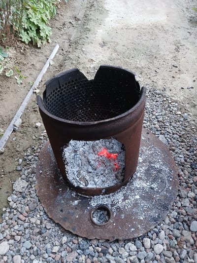 The image depicts a rustic, outdoor scene with a makeshift fire pit. The central object is a rusted, cylindrical metal container, possibly a repurposed drum or pipe, which is open at one end and contains glowing red embers and white ash. The interior of the container shows a perforated surface, suggesting it might have been used for burning or grilling. The cylinder is positioned on a large, flat, circular metal disk, also rusted and covered in ash, which serves as a base or protective layer.
The fire pit is situated on a surface composed of a mix of dry, light-colored soil and small, grey and brown pebbles. To the left of the fire pit, lush green foliage from plants with broad leaves is visible, indicating an outdoor, possibly garden or yard, setting. A thin, metallic strip lies on the ground near the plants and the fire pit.
The scene suggests an activity involving fire, such as cooking or heating, likely taking place during the daytime given the clear lighting. There are no people visible in the image. The overall impression is one of a functional, utilitarian setup in a natural or semi-natural environment. The location context provided is Salar, Uzbekistan, which adds a cultural and geographical dimension to the scene. No text is visible in the image.