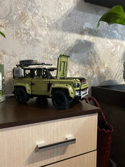 A detailed, forensic, matter-of-fact description of the image:
The image shows a green Lego Technic Land Rover Defender model positioned on the dark brown top of a light-colored wooden dresser with a brushed finish. The dresser has a silver horizontal drawer pull on the visible drawer. The background features a textured wallpaper with a floral pattern in shades of cream and gray. To the left of the model, a green cylindrical object with white stripes and graphics is partially visible. To the right, a dark brown recessed area is present, with a red woven bag inside it. A large green leaf from a houseplant is visible in the upper right portion of the image, casting a shadow on the wallpaper. The lighting suggests an indoor setting with natural light coming from the left. The scene appears to be a still life arrangement on furniture, potentially in a residential environment within the city of Berezovaya Roscha, Russia. There are no people or animals present in the image.