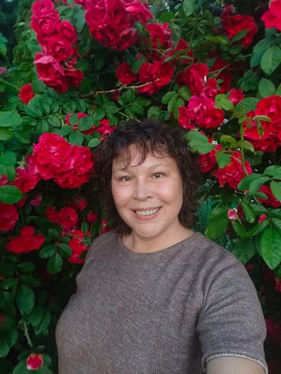A medium shot shows a woman with curly brown hair and a light complexion posing in front of a dense cluster of red roses. She is smiling at the camera, her eyes are open, and her mouth is slightly open. She is wearing a brown, textured shirt with short sleeves. The roses are in full bloom and are a vibrant red color, with green leaves interspersed throughout. The background is dark and out of focus, suggesting the photo was taken outdoors in natural light. The location is Starosemeikino, Russia.
