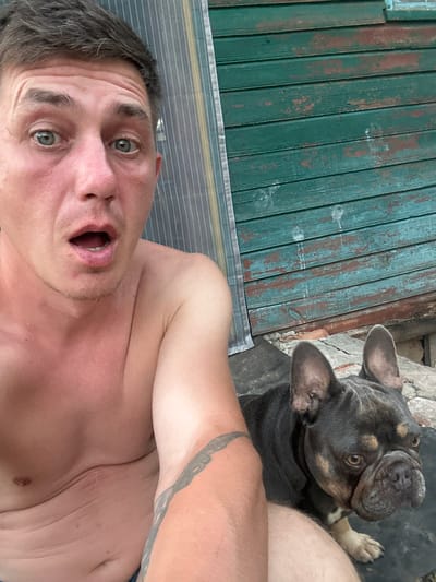 This media file features a selfie taken by a man in Makyshyn, Ukraine, with a French bulldog. The man, positioned in the foreground, appears surprised with his mouth agape and eyes wide open. He is shirtless, revealing his chest and shoulder, with a tattoo visible on his arm. Next to him, a dark-colored French bulldog with distinctive fawn markings is looking upwards. The background shows a weathered green wooden surface and a cracked stone wall, suggesting an outdoor setting. The overall mood is candid and informal, possibly capturing a spontaneous moment between the man and his pet. The time of day is not explicitly indicated, but the lighting suggests it could be daytime.