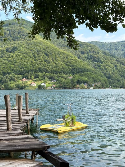 This is a vertical, eye-level shot of a picturesque lake in Ćusine, Bosnia and Herzegovina. In the foreground, a weathered wooden pier juts out into the shimmering blue water, its planks aged and worn. To the right of the pier, a bright yellow, partially submerged boat with green foliage growing on it floats idly, its metallic railings visible. The lake extends to meet a densely forested hillside that dominates the left side of the frame. The trees are a vibrant green, creating a lush carpet that stretches up towards a clear blue sky dotted with a few wispy clouds. Scattered amongst the trees on the hillside are a few small houses with red or grey roofs, suggesting a tranquil, rural settlement. The water is calm with gentle ripples, indicating a peaceful day. The scene evokes a sense of serenity and natural beauty, with the man-made elements of the pier and boat blending harmoniously with the surrounding landscape. There is no visible text in the image. The lighting suggests it is daytime, possibly late morning or early afternoon, with bright sunshine illuminating the scene.