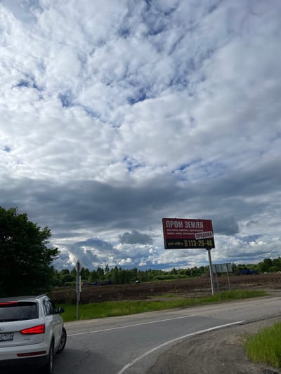 The image shows a roadside scene. In the foreground, a white car is partially visible. Beside the car, a road sign is mounted, displaying an advertisement for land, promoting it for use as a warehouse, parking lot, production, service, car wash, or auto repair shop. The sign prominently displays the phrase "PROM ZEMLYA" (Industrial Land) and provides a phone number (113-26-46) along with the website "park-a108.ru." The background consists of a field and lush greenery, framed by a dramatic sky filled with clouds. According to the license plate, the photo was taken in Ol'khovka, Russia.