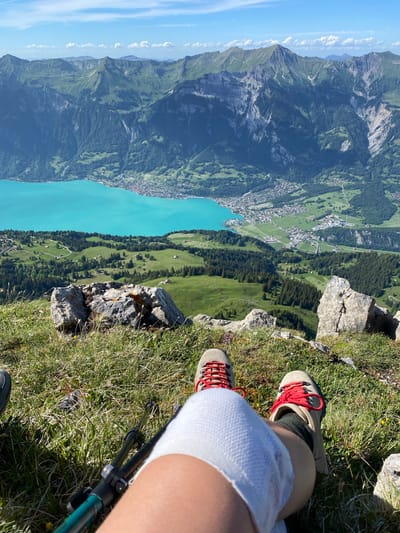 The image captures a scenic view from a mountain peak overlooking the turquoise waters of Lake Brienz and the village of Axalp, Switzerland. In the foreground, a person's legs are visible, wearing hiking boots with red laces and a white bandage around one knee. A hiking pole is partially visible. The ground is covered in green grass and rocky outcrops. In the mid-ground, the lush green slopes of the mountains are dotted with scattered houses and dense pine forests. The background features majestic, rugged mountain ranges under a bright blue sky with scattered white clouds. The overall scene depicts a serene and breathtaking mountain landscape.