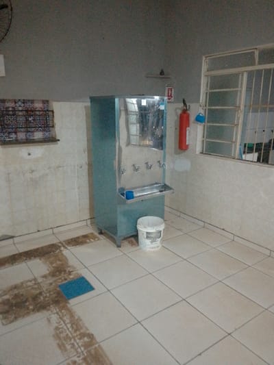 In Cáceres, Brazil, this image captures a utilitarian room. Dominating the center is a teal-colored water cooler with four spigots, positioned on a metal stand with legs. A stainless steel drip tray is attached to the front of the cooler. To the right of the water cooler sits a white plastic bucket with text and graphics visible, its lid slightly ajar. Behind the water cooler, a red fire extinguisher hangs on the wall, with a small blue object, possibly a cup or holder, attached to its side. A window with metal bars is visible above and to the right of the fire extinguisher. The walls are tiled with light-colored square tiles, some showing signs of wear and staining. On the left wall, a decorative tiled panel is mounted, featuring a red, white, and blue geometric pattern. The floor is also tiled with light-colored square tiles, with noticeable darker, stained areas suggesting past spills or wear. A small, bright blue square object, possibly a mat, is placed on the floor in the lower left quadrant. The lighting is dim, creating a somewhat muted atmosphere.