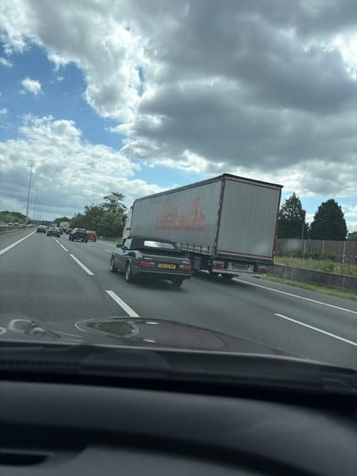 A black convertible Saab is driving on a highway in Ede, Netherlands. The car is in the right lane, and a large white truck is in the left lane. The truck has "Transport Geffels" written on the side of it. The sky is cloudy, and there are trees in the background. The image is taken from inside another car.