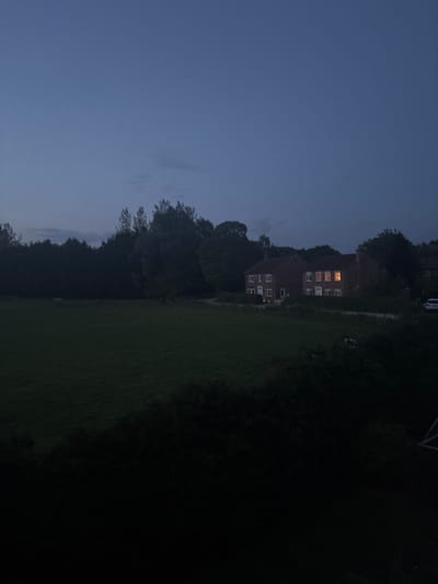 The image shows a single-story brick house with a lit window in the right-most room. The house is situated on a grassy field, with a dark line of trees surrounding it. The sky is a dark blue and the light is fading. The photo was taken from a distance, with the foreground obscured by a thick line of dark foliage. The scene is set in a rural area in Newton upon Derwent, United Kingdom.