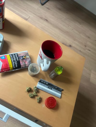 The image displays an overhead view of a light brown wooden surface, likely a table, in an indoor setting located in Herne, Germany. The scene depicts an arrangement of items associated with tobacco, vaping, and cannabis consumption.
In the upper-left corner, a portion of a red container labeled "ZIP CLASSIC PREMIUM TOBACCO" is visible. Adjacent to it, a package featuring a German tobacco warning label is prominently displayed. This label contains a graphic image of severely damaged teeth and gums, accompanied by the German text "Rauchen schädigt Zähne und Zahnfleisch" (Smoking damages teeth and gums) and information for quitting, including "www.rauchfrei-info.de".
Towards the upper right, a white mug with a red interior and a red deer emblem holds a dark liquid. To its right, a light green and silver cylindrical vape device, branded "FLERBAR D6000", rests on the table.
In the lower-center, a small, clear plastic container holds ground green plant material consistent with cannabis. Scattered on the wooden surface nearby are several small, intact buds of green plant material, also consistent with cannabis. A packet of "OCB Premium Slim" rolling papers is positioned next to these buds, alongside a circular, translucent red object identified as a cannabis grinder.
No individuals are present in the frame. The background consists of a wooden floor, and a silver drawer pull is partially visible in the bottom-left corner of the image.