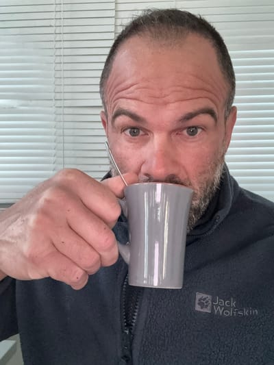 A man is holding a grey mug to his lips. He is wearing a dark grey jacket with a "Jack Wolfskin" logo visible on the upper right chest. The background consists of vertical blinds. The location cannot be determined as Diekirch, Luxembourg based on the image provided.