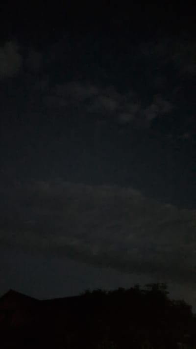 The image captures a dark night sky with scattered clouds over a silhouetted landscape. The lower portion of the image shows the dark outline of rooftops and trees, suggesting an urban or suburban environment. The sky is a deep, dark blue, with lighter grey clouds creating subtle variations in tone. There are no visible people, animals, or specific landmarks that would allow for precise location identification within Hetlingen, Germany, beyond the general indication of a built-up area under a nighttime sky. The overall impression is one of a quiet, possibly cloudy night.