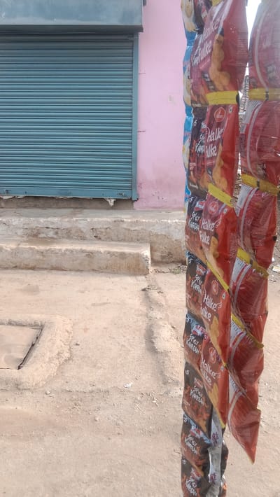The image shows a row of bags of "Spicy Korean Halke Fulke" potato chips hanging from a pole in front of a closed shop with a blue metal roll-up door. The shop is located in Gandhigram, India, at coordinates 23.357 latitude and 80.021 longitude. The bags are arranged vertically, with the top of the pole obscured by the bags. The bags are all red and have a white design featuring a depiction of the chips. The background is a dusty, brown ground, with a concrete step leading up to the shop door.