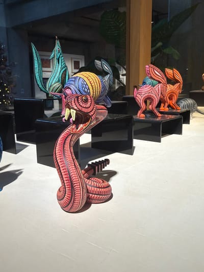 A collection of colorful, hand-painted wooden animal sculptures are displayed on a black, stair-step shelf. The sculptures include rabbits with intricate designs in shades of orange, red, blue, and green. One sculpture depicts a snake-like creature in red tones, coiled and resting on one of the shelf tiers, its shadow cast on the wall. Some sculptures are partially obscured, and there are green plants and reflections in the background. The composition is artistic and visually striking.