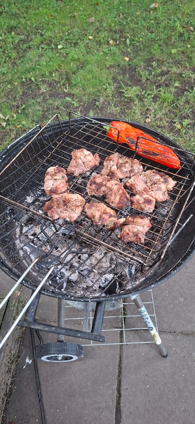 A medium shot captures an outdoor barbecue in progress on a grey paved area, likely a backyard or patio. The barbecue, a circular charcoal grill, is filled with glowing embers and ash, indicating recent use. On the grill rack, several pieces of seasoned meat, appearing to be chicken or pork, are cooking. A whole, grilled red bell pepper lies to the right of the meat, showing signs of charring. The grill itself is black metal with a wire grate and is supported by legs, one of which has a wheel attached. In the background, a patch of green grass and scattered fallen leaves suggest an outdoor, possibly autumnal setting. The image is framed from a high angle, looking down onto the grill.