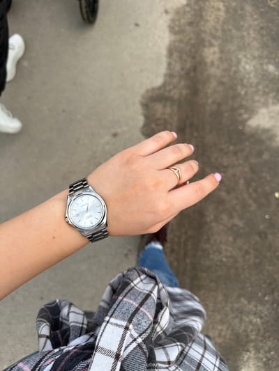 A person is wearing a silver-banded analog wristwatch on their left wrist. The watch has a white face with black hour and minute hands, and a thin red second hand. A date window is visible at the 3 o'clock position. The person is also wearing a gold ring with a small diamond on their right ring finger. The background appears to be an outdoor urban setting, possibly a sidewalk or pavement, with visible texture and some damp patches. The person is wearing blue jeans and a plaid shirt. The overall scene is a close-up shot focusing on the hands and wrist.