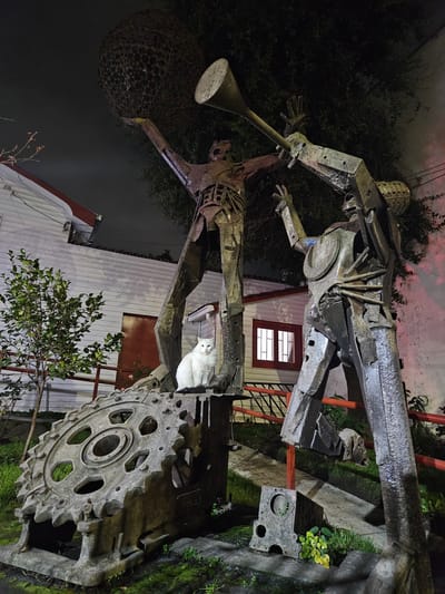 This is a photograph of an outdoor sculpture at night in Chillán, Chile. The sculpture depicts two anthropomorphic figures constructed from scrap metal. The figure on the left is holding a large, spherical, lattice-like object above its head, and the figure on the right is holding a long, conical object that resembles a horn or trumpet. A white cat is perched on a large metal gear, which is part of the base of the sculpture or an associated element. The figures and gear are weathered and appear to be made of salvaged industrial materials. Behind the sculpture is a white building with a red door and a window with red trim. A tree with sparse branches is to the left of the sculpture, and dense foliage is visible behind it and to the right. The ground is covered in grass and moss. The lighting suggests artificial light sources are illuminating the scene.