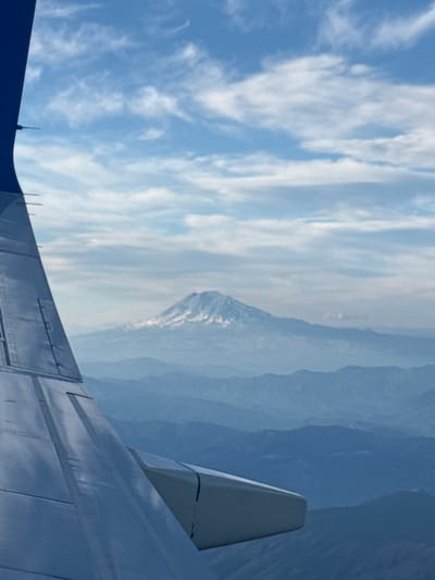 This aerial photograph, taken from an airplane, shows a view of Mount Rainier in the distance. The airplane's wing is visible in the foreground, taking up the left side of the frame. The mountain, capped with snow, stands prominently against a bright blue sky with scattered white clouds. Layered, hazy blue mountains recede into the background, indicating a vast landscape. The specific location in the city of Lost Lake Resort, United States is not discernible from the visual information provided in the image.