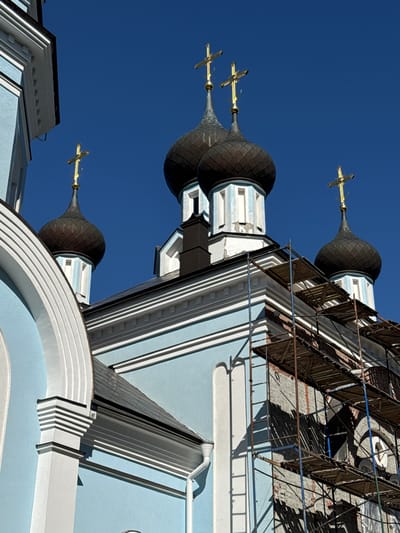 The image shows the exterior of a light blue and white church, likely in St. Ozerki, Russia, under a clear blue sky. Several dark, onion-shaped domes topped with golden crosses are visible. Scaffolding is erected on the right side of the building, suggesting ongoing construction or renovation. The architecture features ornate detailing with white trim against the light blue walls.