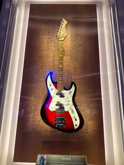 The image shows a signed electric guitar displayed in a glass case. The guitar has a red, black, and white body. It is mounted on a textured bronze-colored wall. There's a plaque to the right of the guitar that reads "KORN". The text describes Korn as one of the first nu-metal groups to break into the mainstream. The scene takes place indoors within a museum-like setting. The environment appears to be lit with recessed lighting at the top. The location of this scene is in the city of Hollywood Seminole Reservation, United States of America.