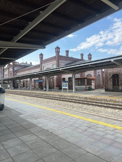 This is a photograph of a train station platform in Kamieniec Ząbkowicki, Poland. The scene is outdoors, under a bright blue sky with scattered white clouds, suggesting daytime with pleasant weather. The platform itself is paved with gray tiles and bordered by a yellow line. Adjacent to the platform are train tracks with gravel ballast.
To the right of the tracks, an old brick building with arched windows and ornate brickwork serves as the station structure. This building is covered in part by a covered walkway supported by arched metal columns, typical of older European train stations. Chimney-like structures with pointed roofs rise from the building's top.
A blue sign is visible on a pole, mounted under the covered walkway, bearing white text. Although slightly blurry, the text appears to read "Kamieniec Ząbkowicki". There are no people clearly visible in the frame. The overall impression is of a historic, functional train station operating on a bright, clear day. The perspective is from the platform, looking along the tracks and towards the station building. A portion of a dark, covered area, possibly another platform or part of the station's roof structure, is visible in the foreground on the left.
