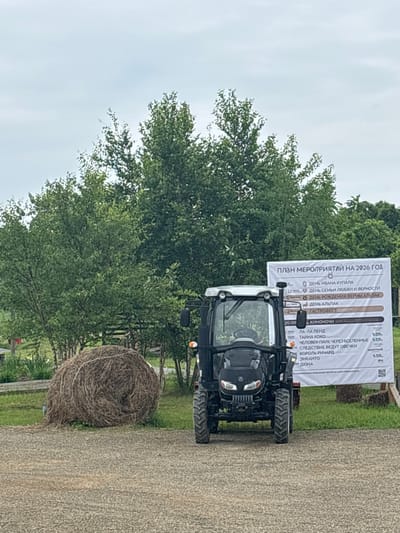 In Tavrichanka, Russia, a front-view shot captures a black tractor parked on a gravel surface. To the left, a large, round bale of hay sits on a grassy area. Behind the tractor, a white banner displays "ПЛАН МЕРОПРИЯТИЙ НА 2026 ГОД" (Plan of Events for 2026) in Russian, along with a list of events. Lush green trees form the backdrop of the scene under a cloudy sky. The image is taken outdoors on a daytime.