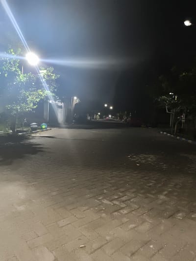 The image depicts a street scene at night in Barengkrajan, Indonesia, illuminated by streetlights. The foreground is a paved walkway made of interlocking bricks. To the left, a leafy tree casts a shadow on the walkway. Two trash bins are visible near the tree. A road extends into the background, lined with more trees and houses. Several streetlights are visible along the road, casting bright beams of light. A red vehicle is parked on the right side of the road in the middle ground. The overall environment appears to be a residential or quiet street.