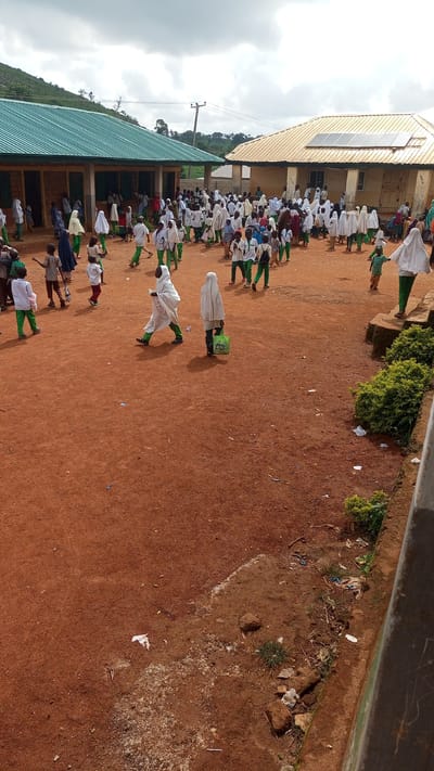 This media file depicts a gathering of people, primarily children, in an outdoor schoolyard. The scene features two buildings with distinct roofing styles: one with a green metal roof and another with a light-colored, possibly tiled roof that has solar panels installed. The ground is covered in reddish-brown soil, suggesting a dry, dusty environment.

The individuals in the image are predominantly dressed in white or light-colored clothing, with many wearing head coverings. Some are wearing green trousers or shirts, forming a contrast. They appear to be engaged in a communal activity or event, possibly a school assembly or a break between classes. The children are spread across the yard, with some walking, others standing in groups, and a few appearing to be interacting with one another. There is a sense of movement and liveliness.

Notable details include the cloudy sky, indicating potentially overcast weather. The presence of the buildings with their different designs, including the solar panels, suggests a rural or semi-rural school setting. The overall atmosphere seems to be one of organized activity, though specific emotions are not clearly discernible from the image. The ground is littered with some debris, including paper and plastic waste, common in such outdoor areas. No visible text is present in the image. The context of Kagarko South, Nigeria, places this scene within a specific geographic and cultural setting.