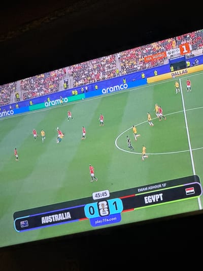 This is a photograph of a large television screen displaying a live soccer match. The game is being played on a green field with white lines marking the boundaries. Several players in red and yellow uniforms are visible on the field. The screen displays a scoreboard at the bottom, showing "AUSTRALIA 0" and "EGYPT 1". The time on the clock reads "45:45". Above the scoreboard, text reads "EMAM ASHOUR 13'". The background of the screen shows a stadium filled with spectators wearing red and yellow clothing. Along the top edge of the screen, banners display "aramco" and "DALLAS". Text in Russian, "НА ЖИВО", is also visible. The overall scene is illuminated by the light of the television screen.