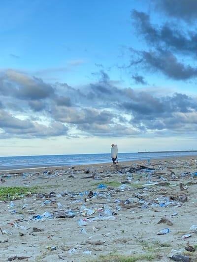 This image depicts a sandy beach in Kualaidi, Indonesia, under a sky with both clear blue areas and scattered dark cumulus clouds. The horizon line is visible, separating the sky from the calm, light blue ocean. Two thin vertical poles are present in the shallow water near the left side of the frame. In the far distance on the right, a structure resembling a pier or breakwater extends into the water.
The sandy foreground of the beach is heavily strewn with an extensive accumulation of various debris, primarily plastic waste. This waste includes numerous white, blue, and transparent plastic bottles, plastic bags, and fragmented plastic items. Interspersed with the plastic debris are pieces of driftwood and sparse patches of green vegetation.
One individual is visible walking on the beach, approximately mid-ground, heading towards the right side of the frame, parallel to the waterline. The person is dressed in dark clothing, wears a light-colored hat, and carries a large, elongated white sack or bag over their left shoulder. The individual is walking on the sand, traversing the littered environment.