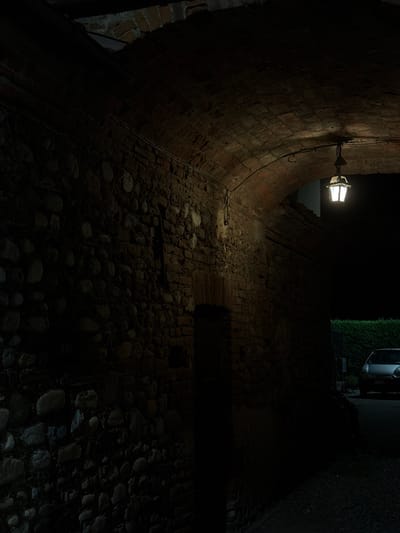 The image shows a dark, narrow alleyway in Lagnasco, Italy, with a brick archway overhead. A vintage street lamp hangs from the archway, casting a warm glow on the cobblestone path and the rough-hewn stone walls. The walls are made of rough stones of various sizes, interlaid with brickwork. The archway is constructed from bricks. On the right side of the alley, a small silver car is parked. A dense green hedge lines the far end of the alley, and a metal gate is visible. The scene is dimly lit, suggesting it is nighttime.