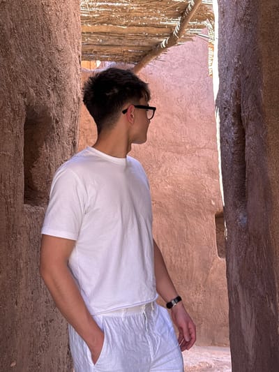 The scene depicts a male individual standing within a narrow, sunlit passageway constructed of reddish-brown adobe or mudbrick, characteristic of the Ouarzazate region in Morocco. The ceiling comprises crude wooden beams supporting a layer of thatch or reeds, allowing sunlight to penetrate. The individual, seen from the mid-torso, faces right in profile, wearing black-framed glasses, a white short-sleeved t-shirt, and white linen trousers. His right hand is inserted into a trouser pocket. A black wristband with a silver-toned clasp is on his left wrist. The wall to the viewer's left features a small, rectangular alcove.