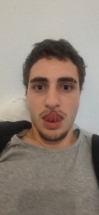 A young man with dark hair and brown eyes is sticking out his tongue at the camera. He is wearing a gray t-shirt and has a short beard. The background is a plain white wall. His face is centered and takes up most of the frame. The image appears to be a selfie taken indoors.