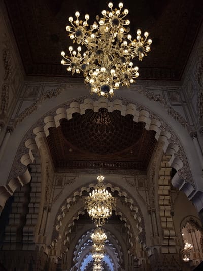 The image displays the interior of a large, ornate architectural structure, located in Bouznika, Morocco. The scene is dominated by a series of grand, illuminated chandeliers suspended from a high, dark, intricately patterned ceiling. The largest chandelier, centrally positioned in the upper frame, is a multi-tiered fixture with numerous radial arms, each terminating in a glowing light bulb. Below it, a linear sequence of progressively smaller, similar chandeliers recedes into the background, forming a luminous path along the central axis of the hall.

The architectural elements consist of multiple pointed arches that extend into the depth of the structure. These arches and the surrounding walls are extensively decorated with elaborate white or light-colored geometric and curvilinear patterns, likely carved or stuccoed, contrasting against darker recessed areas. The ceiling above the arches also exhibits dark, complex decorative motifs. No individuals or specific actions are depicted within the frame. The primary light source is artificial, originating from the illuminated chandeliers, which cast a warm glow throughout the richly adorned interior.