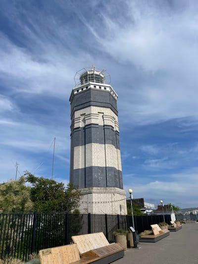 The image shows a tall, striped lighthouse with alternating bands of white and dark gray brick. The lighthouse is viewed from a low angle, looking up towards the sky, which is filled with wispy white clouds against a bright blue background. The lighthouse has a metal railing around the top observation deck.
In the foreground and to the right of the lighthouse, there is a paved walkway with several modern wooden benches and planters with greenery. A black metal fence runs along the edge of the walkway. Beyond the lighthouse and walkway, buildings and distant hills are visible, suggesting an urban or coastal setting.
The lighting suggests it is daytime, with bright sunlight casting shadows. The sky appears clear and partly cloudy, indicating fair weather. There are no people visible in the image. The overall scene depicts an outdoor public space featuring a prominent lighthouse. Visible text is limited to a small logo on one of the wooden benches. The location context is Varvarovka, Russia.
