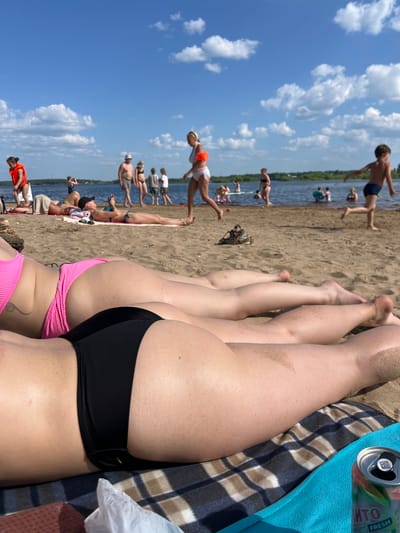 On a sunny day at the beach in Ust'ye, Russia, several people are enjoying the water and sand. In the foreground, two women are lying on a patterned blanket. The woman on the left wears a pink bikini, while the woman on the right wears black bikini bottoms. Their legs are sunbathing on the sandy beach. In the background, other beachgoers are visible. Some are in the water, while others are walking along the shore or lying on towels. A young boy in blue shorts runs towards the water. The sky is blue with scattered white clouds, and the water appears calm. A can of what appears to be a beverage is placed on the blanket in the lower right corner of the image. The environment suggests a leisurely summer day at a lakeside or river beach.